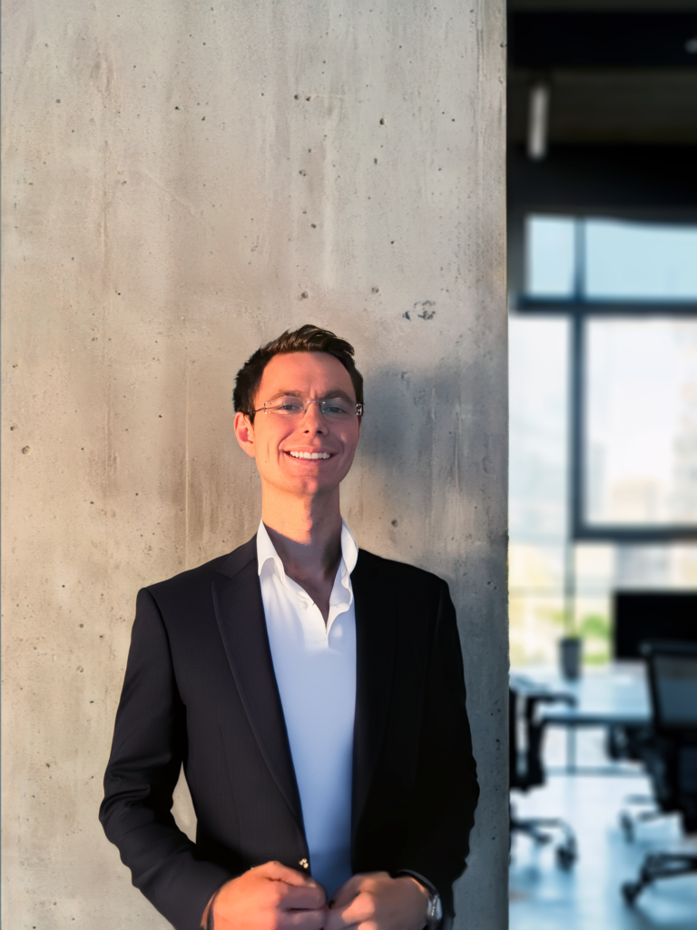 Professional headshot of Grayson Mylar in a modern office setting with concrete wall and glass windows. He wears a navy blazer over a white shirt, standing confidently with natural lighting and a blurred office background